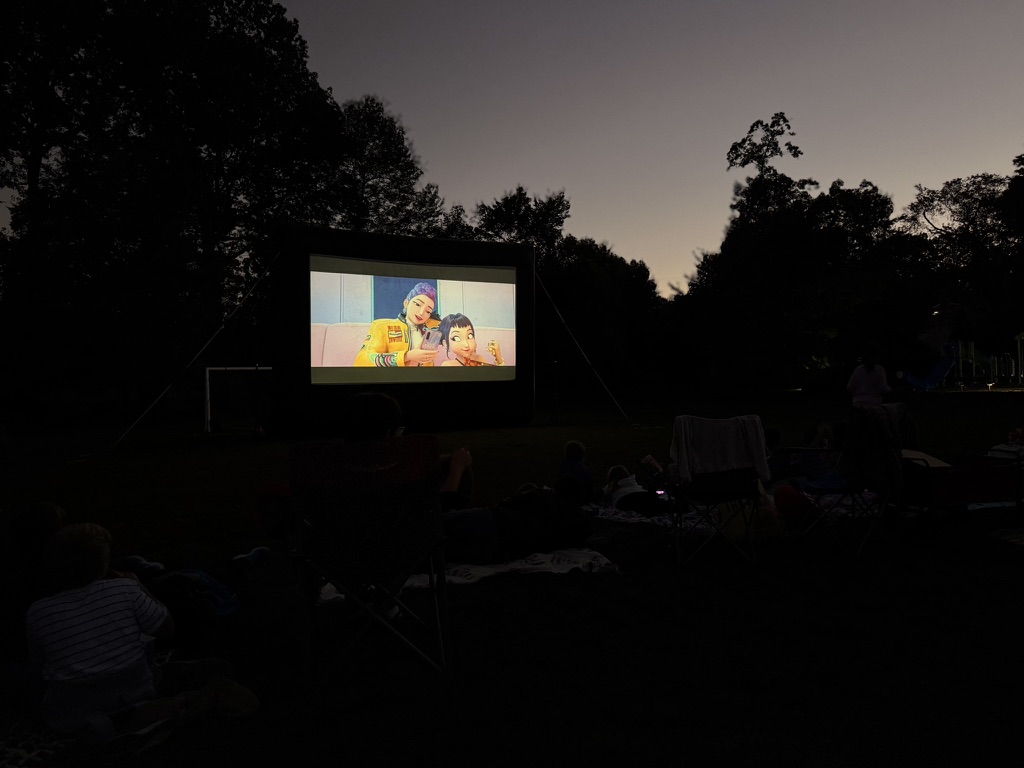 Families on blankets and lawn chairs watching a movie on a big outdoor screen at dusk, last year's Film on the Field
