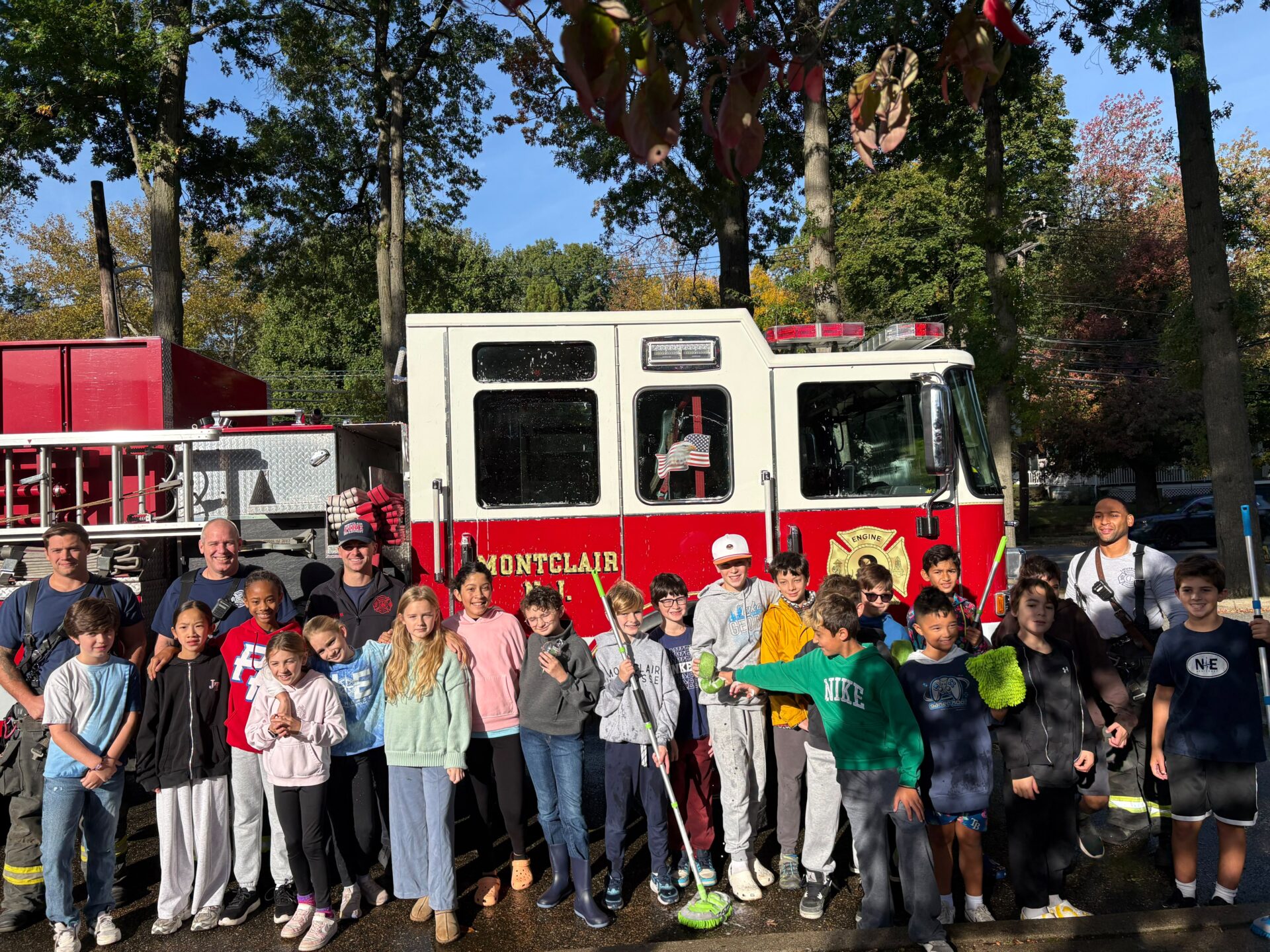 Northeast 5th graders holding car-wash brushes in front of a Montclair fire engine, with four Montclair firefighters, at the Fifth Grade Car Wash